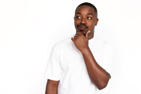 Pensive African American man touching chin. Portrait of unsure young male model with short hair in white T-shirt looking away, pouting lips with hand on chin. Advertisement, contemplation conceptの写真素材