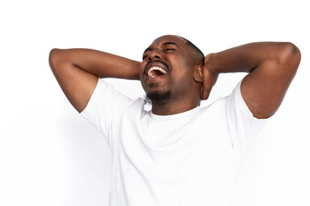 Desperate African American man screaming with hands on head. Portrait of shocked young male model with short hair and beard in white T-shirt with closed eyes suffering from headache. Despair conceptの写真素材