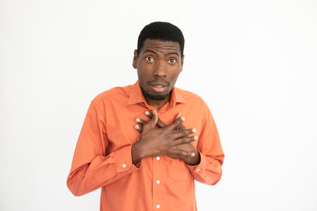 Portrait of ashamed young man touching chest over white background. African American guy wearing orange T-shirt making excuses. Apology conceptの写真素材