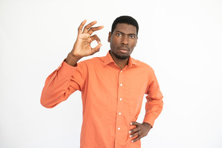 Portrait of confident young man making okay gesture over white background. African American guy wearing orange T-shirt looking at camera with serious expression. Success and satisfaction conceptの写真素材
