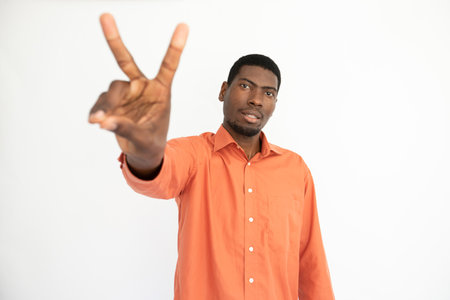 Portrait of happy young man making victory gesture over white background. African American guy wearing orange T-shirt showing peace sign and smiling at camera. Fun and happiness conceptの写真素材