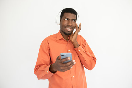 Portrait of candid young man listening to mobile player in headphones. African American guy wearing orange shirt using music app on smartphone. Music and mobile technology conceptの写真素材