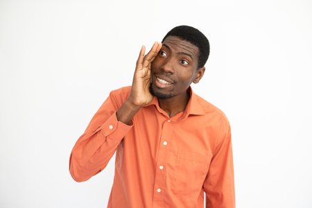 Portrait of happy young man telling gossip covering mouth with hand. African American guy wearing orange T-shirt looking away and smiling. Secret and gossip conceptの写真素材