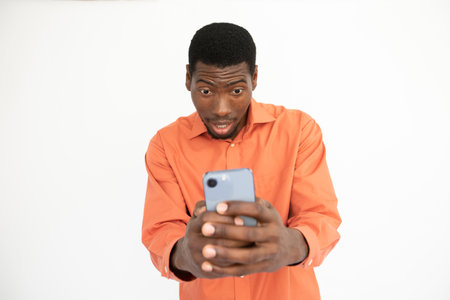 Portrait of astonished young man reading newsletter on phone. African American guy wearing orange shirt surfing internet or reading message. Mobile technology and astonishment conceptの写真素材
