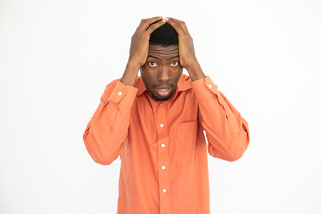 Portrait of frustrated young man holding head in hands over white background. African American guy wearing orange shirt looking at camera in fear. Problem or trouble conceptの写真素材