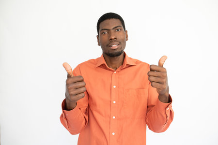 Portrait of positive young man making thumb up gesture over white background. African American guy wearing orange T-shirt looking at camera and smiling. Approval conceptの写真素材