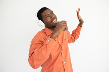 Portrait of young man listening to music and recording voice. African American guy in headphones wearing orange shirt singing over white background. Music and mobile technology conceptの写真素材