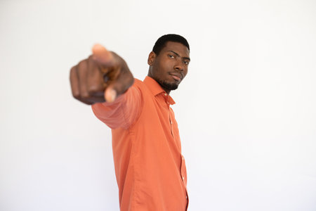 Portrait of confident young man pointing at camera over white background. African American guy wearing orange shirt blaming or hiring something. Blame or recruitment conceptの写真素材