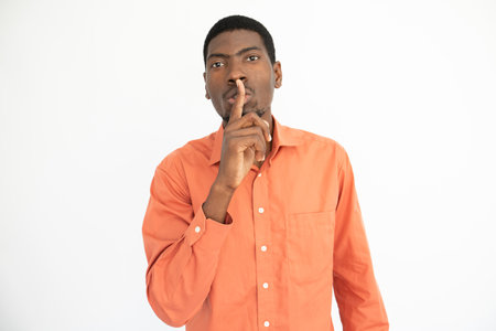 Portrait of confident young man posing with finger on lips over white background. African American guy wearing orange shirt showing silence gesture. Secret and silence conceptの写真素材