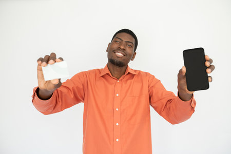 Portrait of happy young man showing mobile phone and credit card over white background. African American guy wearing orange shirt advertising contactless payment. Banking conceptの写真素材