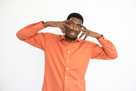 Portrait of happy young man holding fingers at eyes over white background. African American guy wearing orange T-shirt looking at camera and smiling. Fun and happiness conceptの写真素材