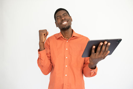 Portrait of successful young man making winning gesture. African American guy wearing orange shirt holding digital tablet and celebrating success. Wireless technology and success conceptの写真素材