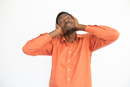 Portrait of happy young man covering ears with hands over white background. African American guy wearing orange T-shirt looking at camera and smiling. Ignorance conceptの写真素材