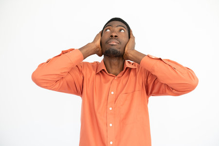 Portrait of ignorant young man covering ears with hands over white background. African American guy wearing orange T-shirt protecting ears from noise. Ignorance conceptの写真素材