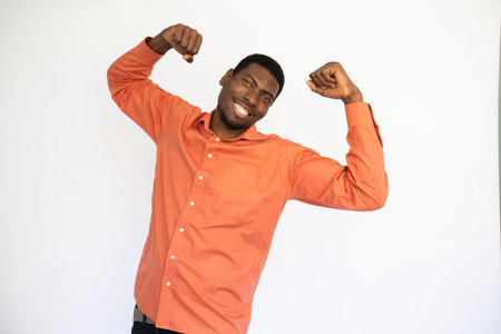 Portrait of happy young man showing biceps over white background. African American guy wearing orange shirt expressing strength. Fitness and power conceptの写真素材
