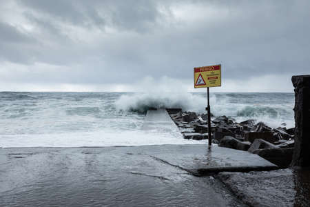 MADEIRA, PORTUGAL - NOVEMBER 22, 2017: danger warning sign in a marina on a stormy day with big waves hitting the coastの写真素材