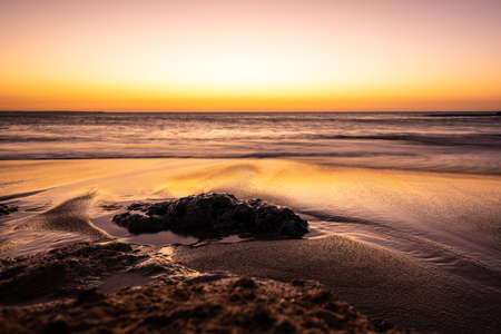 long exposure photo of swirl pattern of waves rolling onto sand beach with scattered rocks at sunset...