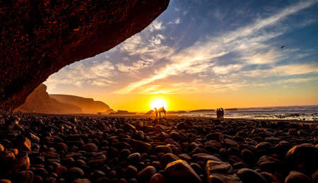sunset sky over natural phenomenon stone arch bridge of Legzira Beach with dromedary camel in front...