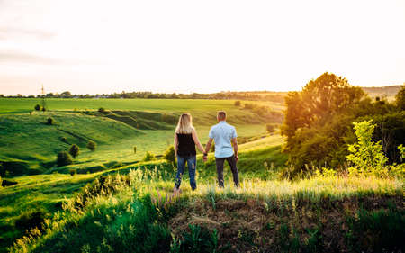 Girl and guy holding hands and standing on a green hill. Sunset. Back view.の写真素材