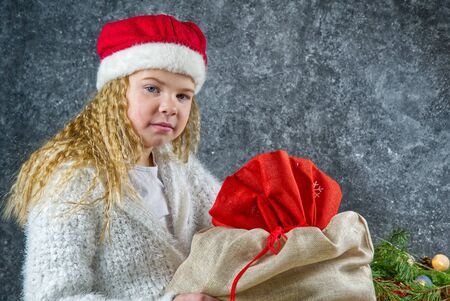 Little blonde girl in a red gnome hat unhappy with a bag of gifts from Santa in anticipation of a Christmas miracle.の写真素材