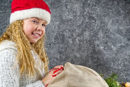 Little blonde girl in a red gnome hat Happy with a bag of gifts from Santa in anticipation of a Christmas miracle.の写真素材