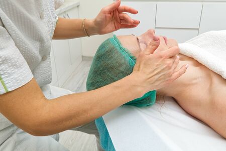Beautician applies a natural moisturizing mask to the face of a young beautiful woman. Anti-aging mask and wrinkles for problem skin. Cosmetology concept. The cosmetologist performs the procedure to cleanse and moisturize the face.の写真素材