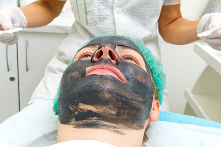 Young woman in a beauty salon with a black mask on her face. The concept of cosmetology. The cosmetologist performs a procedure to cleanse the face with problem skin.の写真素材