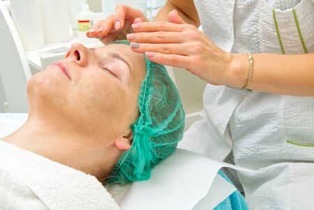 Beautician applies a natural moisturizing mask to the face of a young beautiful woman. Anti-aging mask and wrinkles for problem skin. Cosmetology concept. The cosmetologist performs the procedure to cleanse and moisturize the face.の写真素材