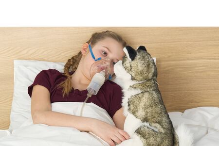 girl with inhaler mask - respiratory problems for asthma. girl with a mask inhaler lies with a toy in bed and breathes adrenaline. health care and sick child conceptの写真素材