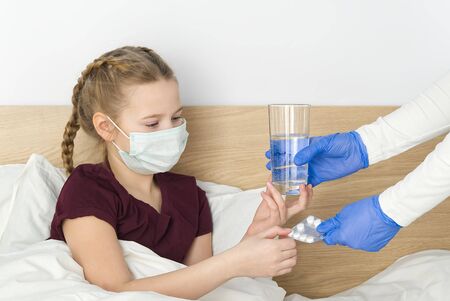 A young girl lying on the bed is given pills and a glass of water. masked girl for protection against viral diseases. concept of quarantine and treatment for viral diseases, colds and coronavirusの写真素材