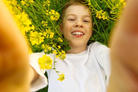 Happy smiling boy making self portrait on smartphone in meadow. young boy making selfie on smartphone laying in green grass with yellow flowers.の写真素材
