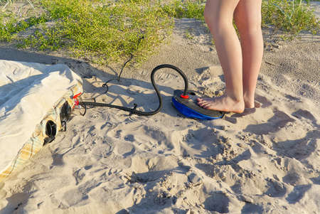 A woman with air foot pump pumps an inflatable mattress or air bed at sandy beach. Foot inflates air mattress with foot pump on sand.の写真素材