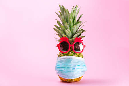 COVID Travel concept. Pineapple in sunglasses and medical mask on a pink background. Travel or Vacation idea design.の写真素材