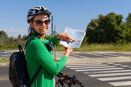 tourist, happy woman in a helmet on a bicycle examining a map at the intersection of bicycle roads. concept of outdoor activities or cycling travel in sunny dayの写真素材