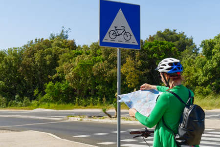 tourist, happy woman in a helmet on a bicycle examining a map at the intersection of bicycle roads. concept of outdoor activities or cycling travel in sunny dayの写真素材