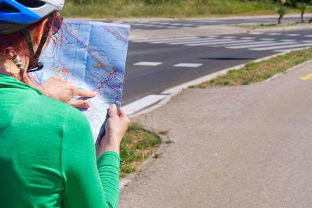 tourist, happy woman in a helmet on a bicycle examining a map at the intersection of bicycle roads. concept of outdoor activities or cycling travel in sunny dayの写真素材