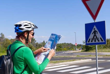 tourist, happy woman in a helmet on a bicycle examining a map at the intersection of bicycle roads. concept of outdoor activities or cycling travel in sunny dayの写真素材