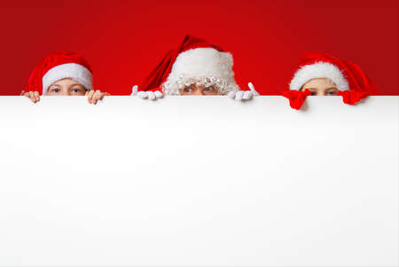 satna claus with funny children in red christmas gnome hats peeking out from behind a blank white banner, mockup and copy spaceの写真素材