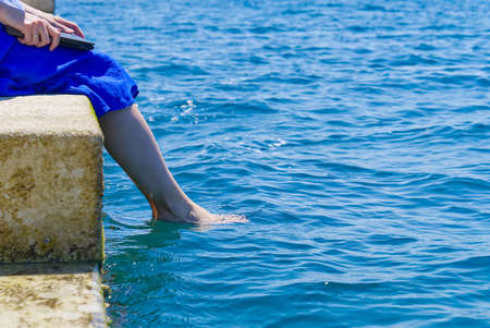 Work from anywhere. Side view of young woman, female freelancer in straw hat working on laptop while sitting on the beach. young woman in sitting on stone at sea and remotely working on laptopの写真素材