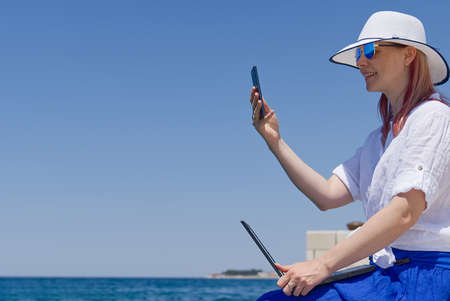 Work from anywhere. Side view of young woman, female freelancer in straw hat working on laptop while sitting on the beach. young woman in sitting on stone at sea and remotely working on laptopの写真素材