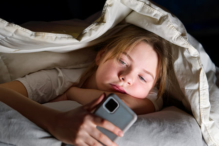 Gadget night. Romantic chat. Late home leisure. Online communication. Portrait of girl with mobile phone under blanket at night. cyber bullyingの写真素材