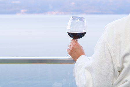The elegant female hand holding a glass of red wine with the background of a sunny seaside view.の写真素材