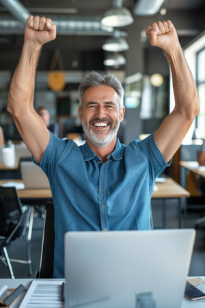 Senior business professional celebrating success in front of laptop with arms raised in joyの素材