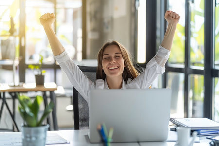 Successful businesswoman celebrating victory in front of laptop with arms raised in joyの素材