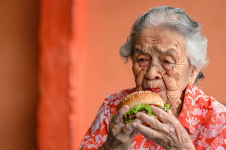 Senior woman enjoying tasty burger on soft colored backdrop, ideal for text placementの素材