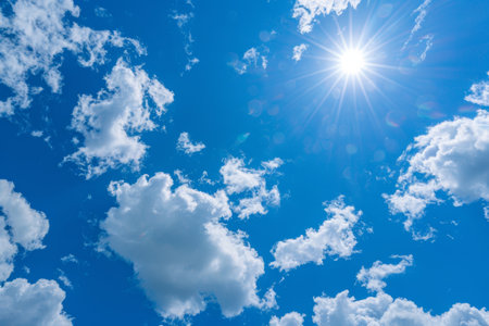 Bright sun shining through fluffy white clouds on a vibrant blue sky, sunny weather backgroundの素材