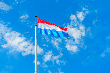 Flag of Luxembourg waving against a clear blue sky with white clouds on a sunny day