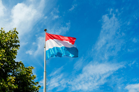 Flag of Luxembourg blowing in the wind against a blue sky with white cloudsの写真素材