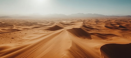 Vast, orange sand dunes under a bright sun, with mountains on the horizon. Desert landscape.の素材