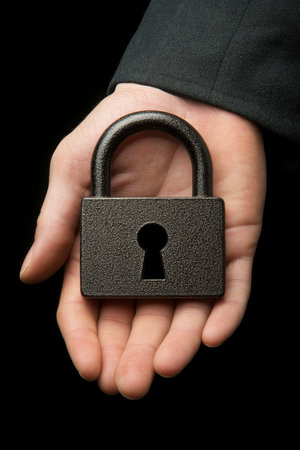 Close-up of a businessmans hand securely holding a black padlock, isolated on a black background.の素材